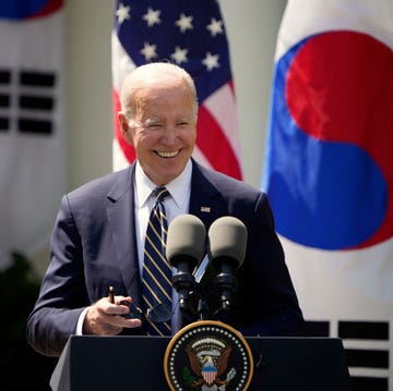 joe biden, wearing a dark blue suit jacket, white shirt, and striped tie, smiles while standing at a podium with two microphones on it, with american and south korean flags behind him