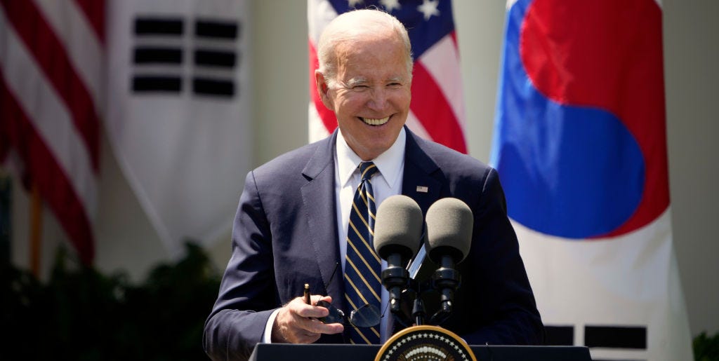 President Joe Biden at The White House joe biden, wearing a dark blue suit jacket, white shirt, and striped tie, smiles while standing at a podium with two microphones on it, with american and south korean flags behind him