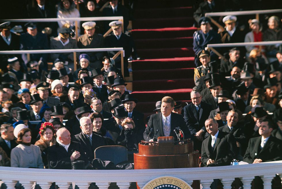 john f kennedy speaks as he stands behind a wooden podium on a balcony, a crowd of people sits behind him and watches