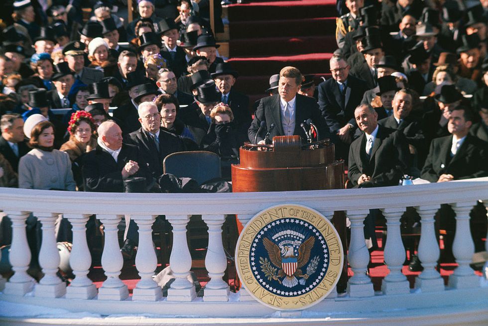 john f kennedy speaks as he stands behind a wooden podium on a balcony, a crowd of people sits behind him and watches