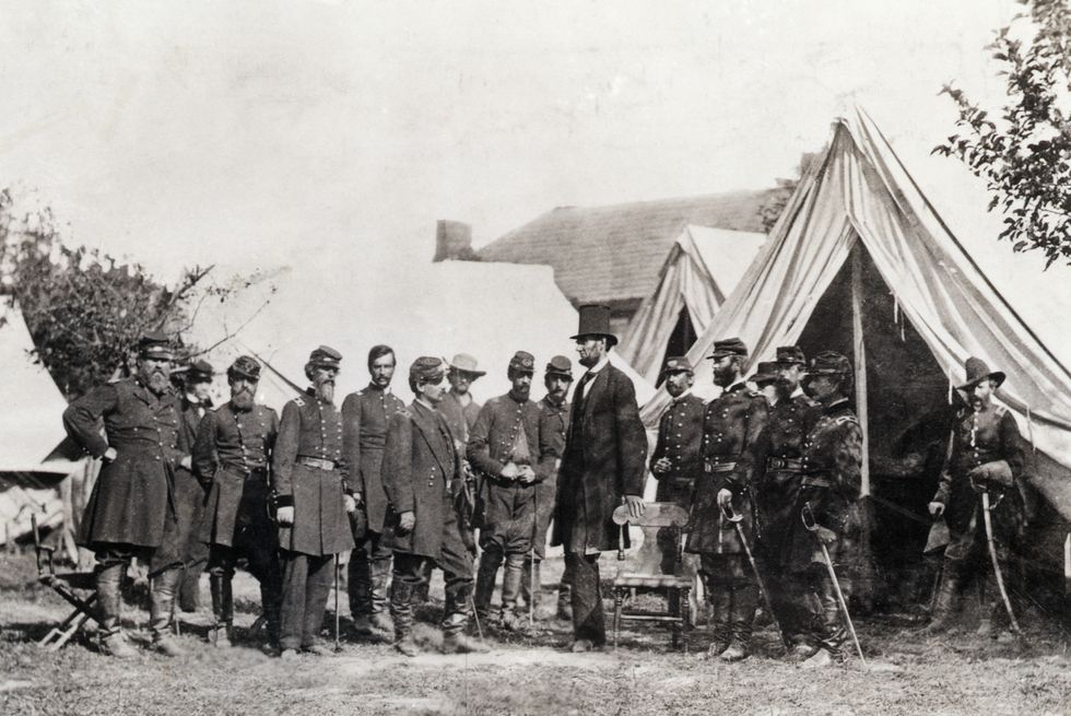 abraham lincoln stands next to 15 union army soldiers in uniform at a war camp, lincoln holds onto the back of a chair and wears a long jacket and top hat