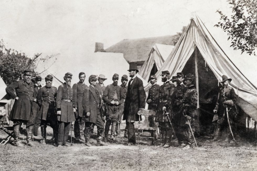 abraham lincoln stands next to 15 union army soldiers in uniform at a war camp, lincoln holds onto the back of a chair and wears a long jacket and top hat