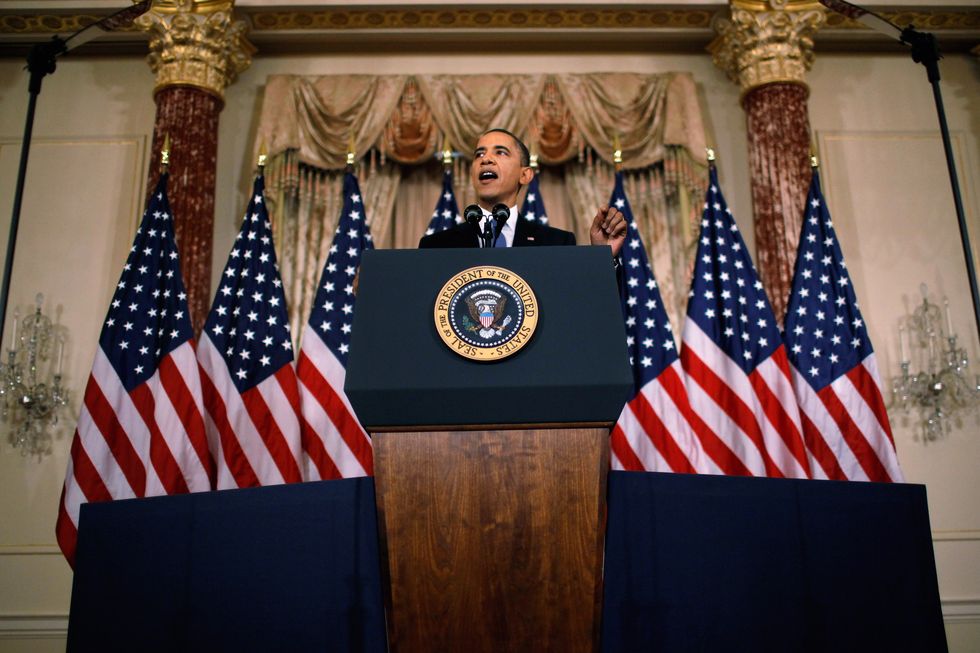 barack obama delivers a speech at a podium outfitted with the presidential seal of the united states, behind him are eight american flags and an ornate room with two columns, two chandeliers, and a large window with curtains