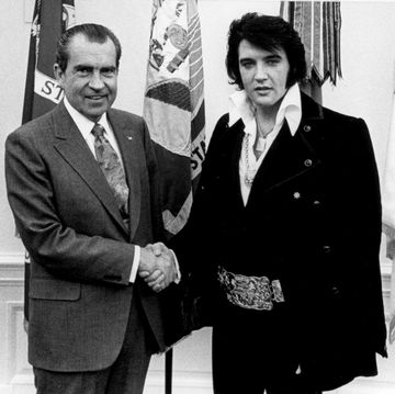 two men in suits shake hands while posing for a photo in front of several flags