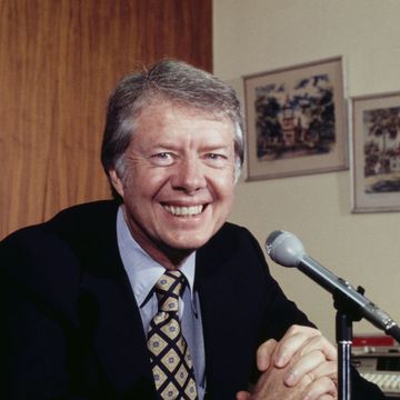 jimmy carter smiles at the camera while seated behind a microphone, two framed paintings hang on a wall behind