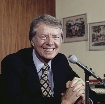 jimmy carter smiles at the camera while seated behind a microphone, two framed paintings hang on a wall behind