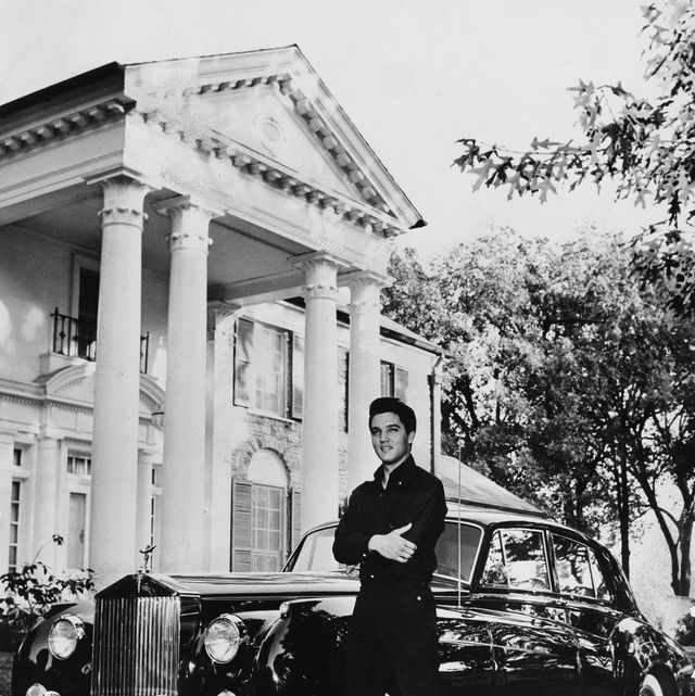 a man stands with his arms crossed and smiling, behind him is a black car and a large house with four pillars