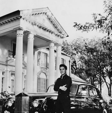 a man stands with his arms crossed and smiling, behind him is a black car and a large house with four pillars