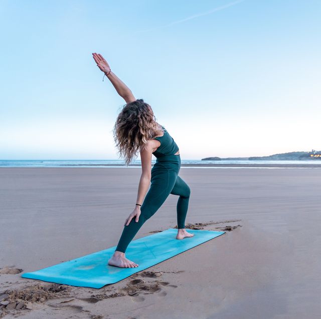 pretty blonde woman doing yoga in front of the sea on the beach meditating on blue mat at sunrise