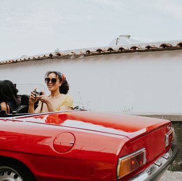 a pretty young woman sits in the back of a vintage red convertible and takes photos with a retro film camera