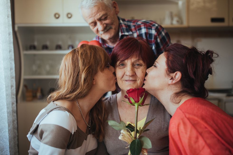 two grownup daughters kissing their mom's cheeks, mom holds a rose, elderly father stands behind them smiling