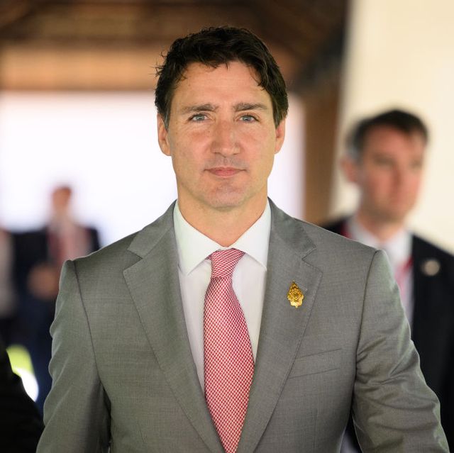 justin trudeau looks past the camera while standing in a room, he wears a gray suit jacket, white collared shirt and red and white tie with a gold lapel pin