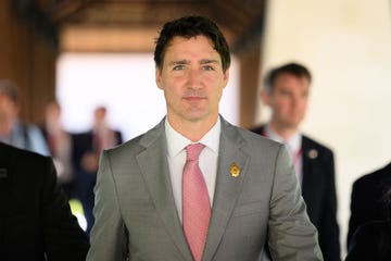 justin trudeau looks past the camera while standing in a room, he wears a gray suit jacket, white collared shirt and red and white tie with a gold lapel pin