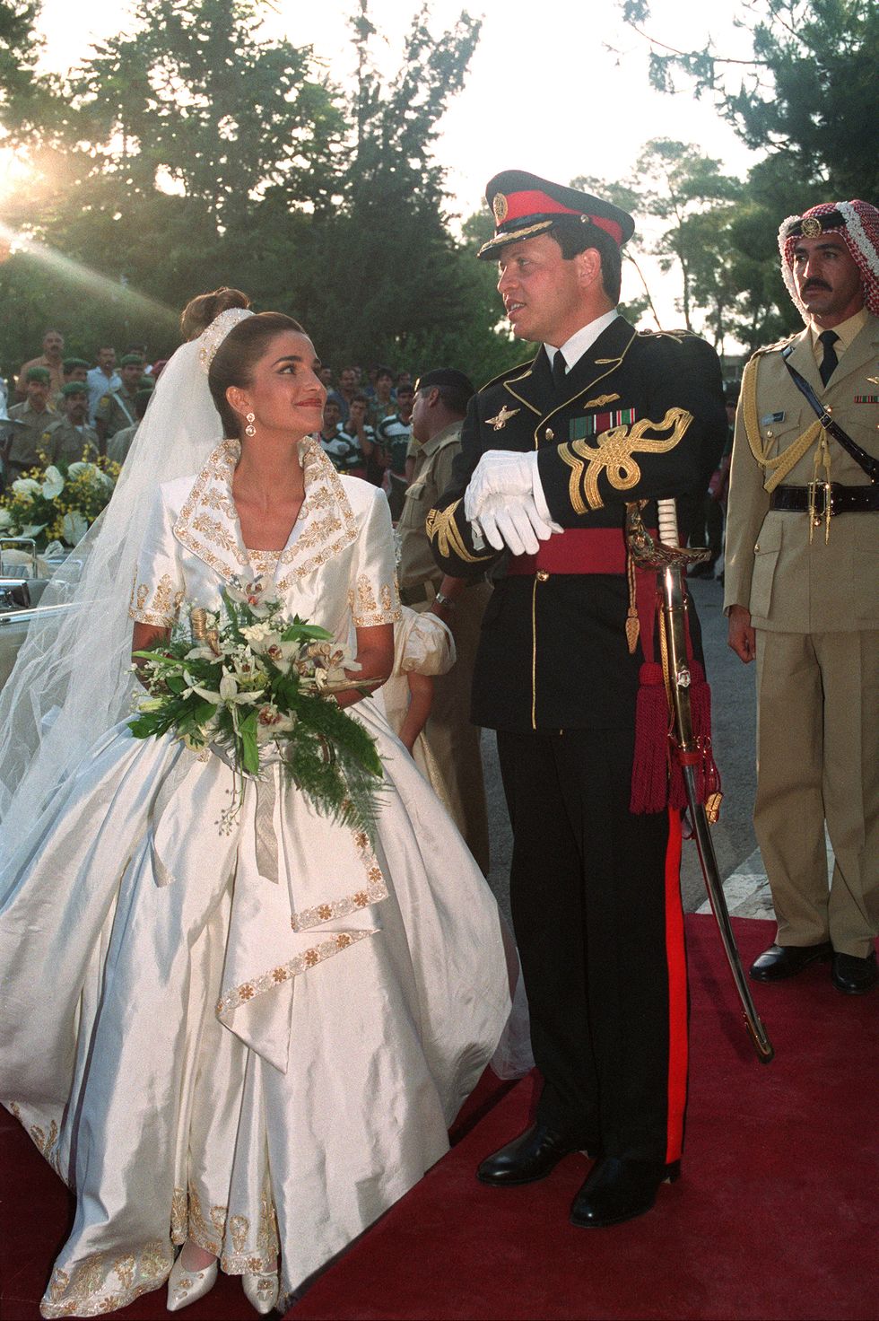 rania al yassin stands on a red carpet outside next to prince abdullah ii, she looks at him smiling and wearing an ornate ballgown style wedding dress with a long veil, she also holds a bouquet of greenery and white flowers, he wears a black military uniform with white gloves and a hat