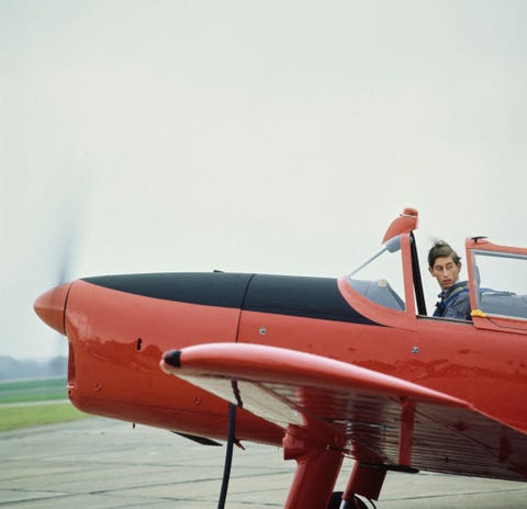 prince charles sits in the pilot seat of a red and black propeller plane and looks over his shoulder