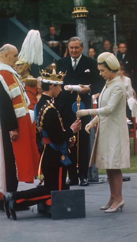 prince charles kneels before queen elizabeth ii who is standing in front of him and passing him a golden staff, charles is wearing a decorated military uniform and crown, elizabeth is wearing a yellow outfit and short tan heels