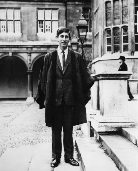 prince charles stands in a courtyard on a set of steps while wearing academic robes and a suit, he is smiling at the camera