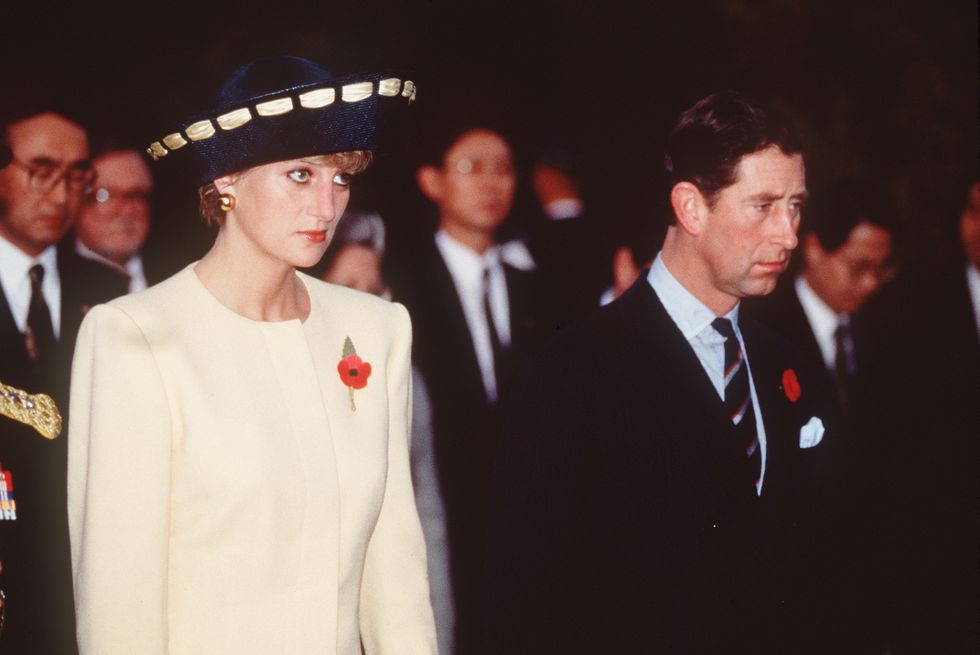 princess diana, wearing a white pantsuit and blue hat, standing next to prince charles, wearing a black suit and blue tie, both unsmiling, with several people in suits behind them