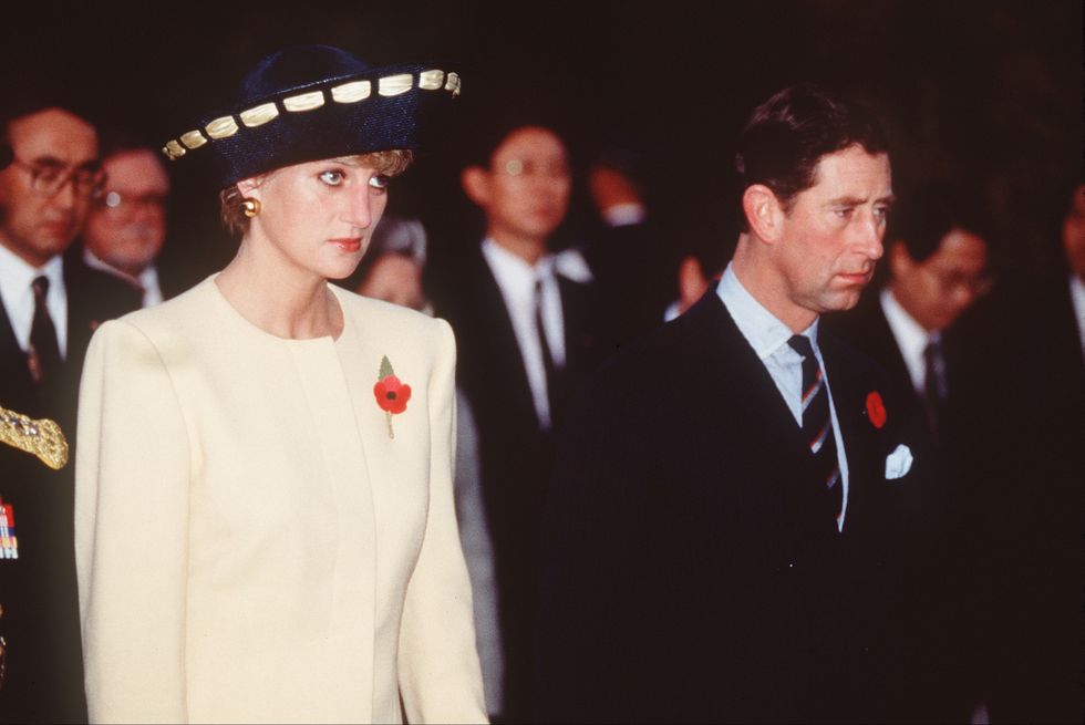 princess diana, wearing a white pantsuit and blue hat, standing next to prince charles, wearing a black suit and blue tie, both unsmiling, with several people in suits behind them