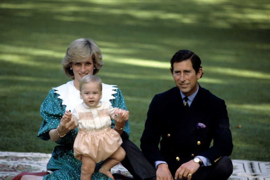 princess diana and prince charles smile while seated on a blanket on a lawn, diana helps prince william as a baby stand by holding both his hands in hers