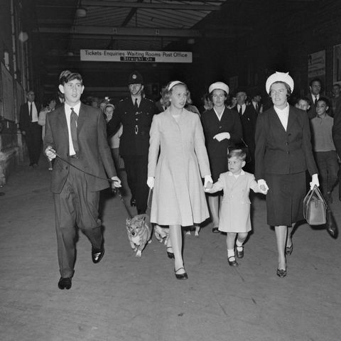 prince charles, princess anne, and prince andrew walk at a train station, charles is walking a corgi and andrew is holding the hand of a nanny, behind them are other stationgoers