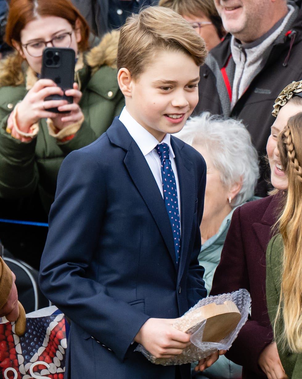 Prince George of Wales on December 25, 2023 prince george of wales smiles as he stands next to people, he holds a wooden plaque in his hands and wears a blue suit with a white collared shirt and blue patterned tie