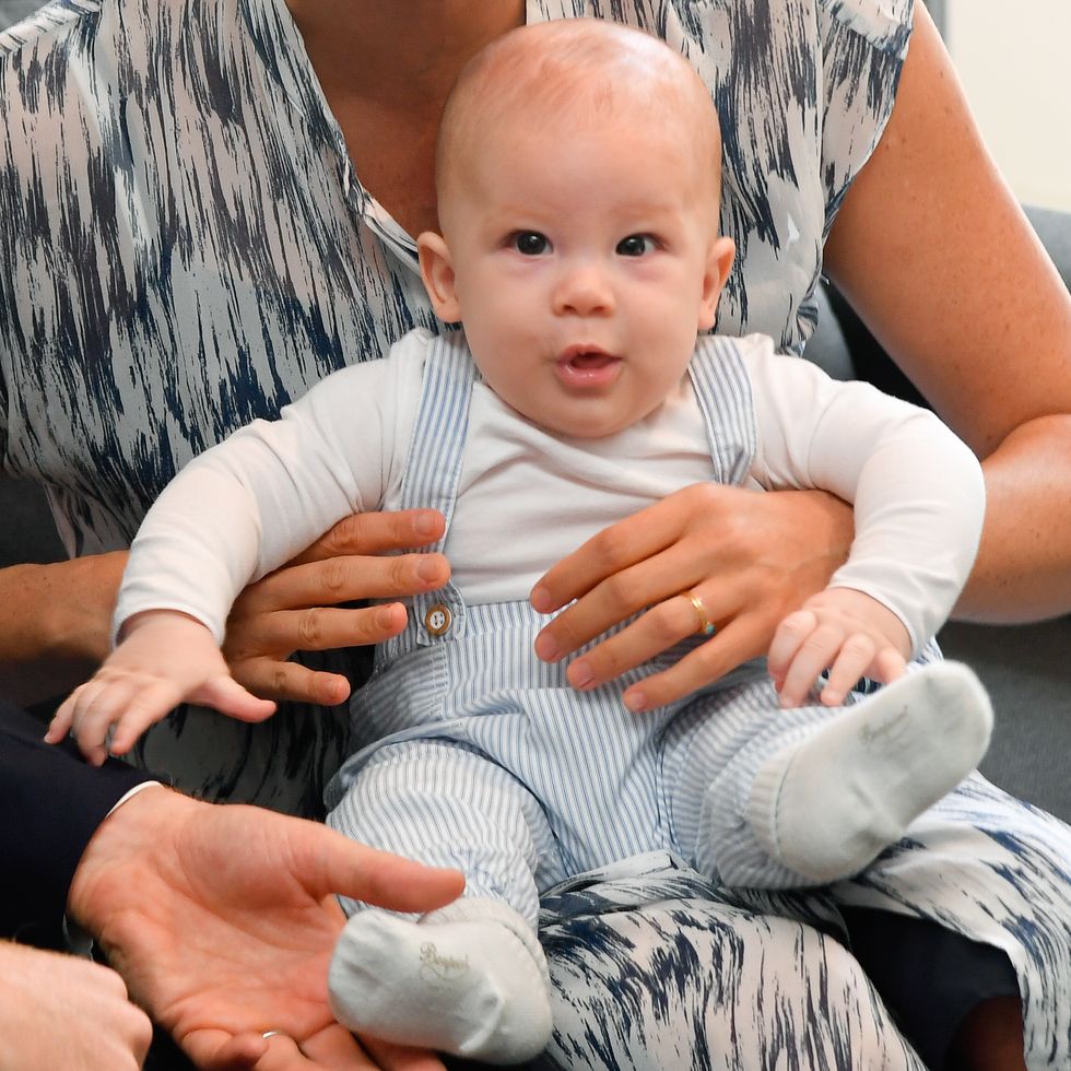 archie mountbatten windsor as a four month old baby sits on a woman's lap, he is wearing a white long sleeve shirt, a blue and white striped jumper and white socks, he is looking at the camera as the woman's hands close around his chest to support him