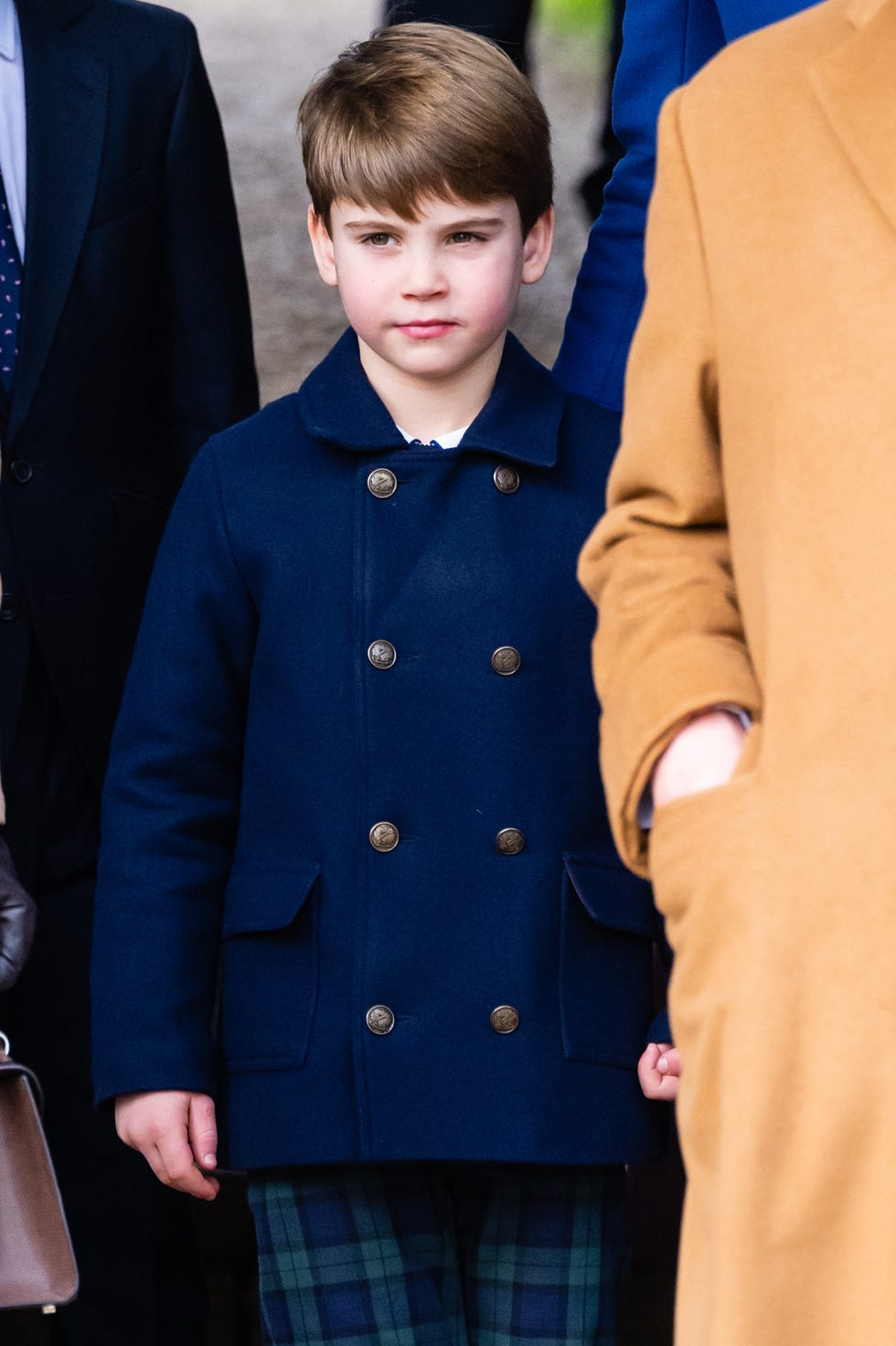 Prince Louis of Wales on December 25, 2023 prince louis of wales stands in a blue peacoat and blue and green plaid pants among other people