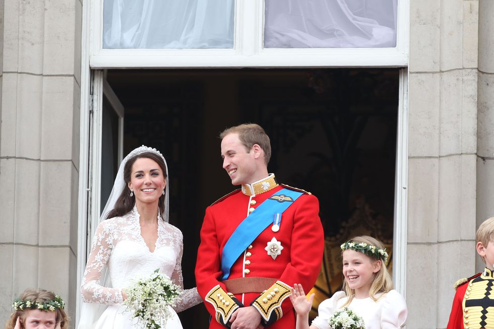 princess kate and prince william stand on a balcony and smile, she wears a white lace wedding dress and holds a bouquet, he wears a red military uniform with a blue sash, two young girls stand nearby wearing flower crowns and dresses