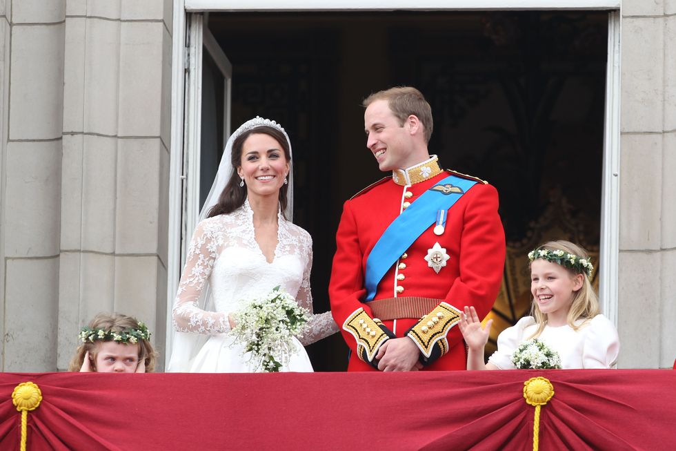 princess kate and prince william stand on a balcony and smile, she wears a white lace wedding dress and holds a bouquet, he wears a red military uniform with a blue sash, two young girls stand nearby wearing flower crowns and dresses