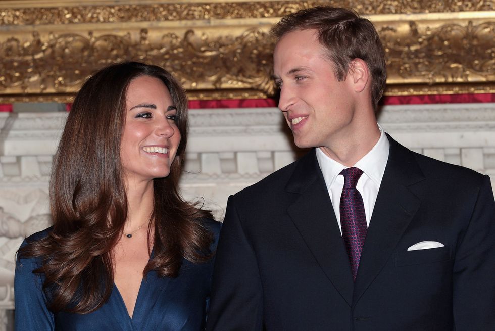kate middleton and prince william look at each other and smile, she wears a blue dress, he wears a black suit jacket with a white collared shirt and red and navy tie