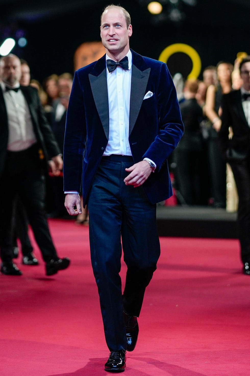 Prince William of Wales attends the 2024 BAFTA Film Awards in February prince william walks on a red carpet while looking slightly up and smiling, he wears a blue velget suit with a bow tie and white collared shirt