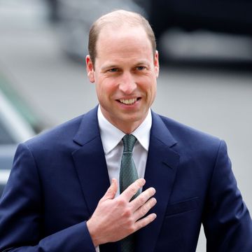prince william smiles he walks outside, he holds one hand close to his chest and wears a navy suit jacket, white collared shirt and green tie