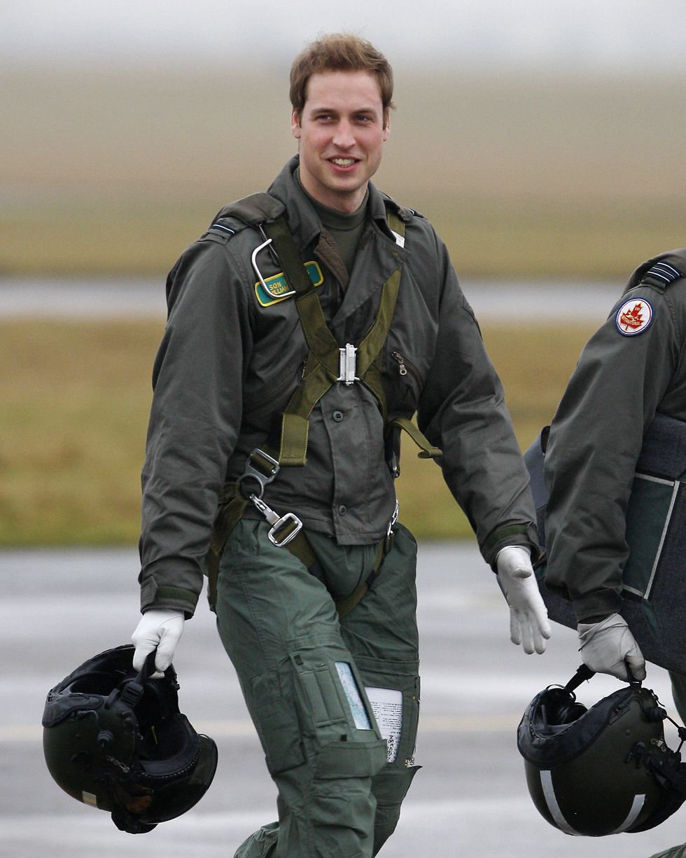 prince william walks across tarmac outside while wearing military fatigues, a harness and white gloves, he holds a helmet in one hand