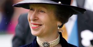 princess anne smiles and looks left, she wears a hat and blue velvet jacket over a white and blue top with brooches and pins, she also has on a pearl necklace and earrings