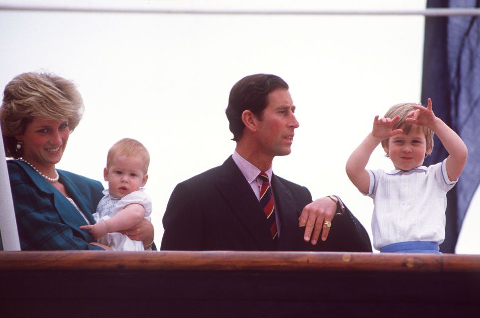princess diana holds baby ﻿prince harry and stands next to then prince﻿ charles and prince william behind a wooden railing