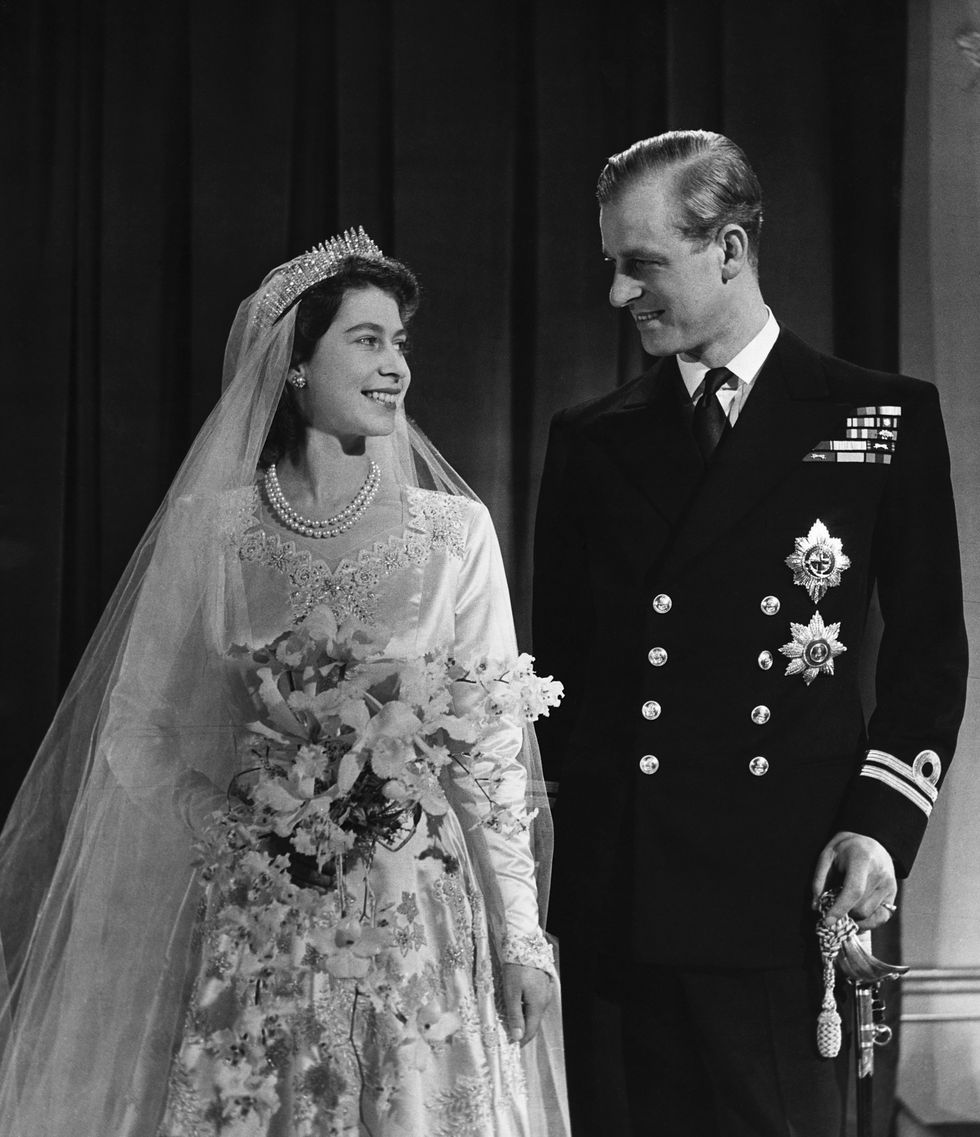princess elizabeth and philip mountbatten stand and look at each other smiling, she wears a wedding dress, veil and crown and holds a bouquet, he wears a dark military uniform and holds a sword