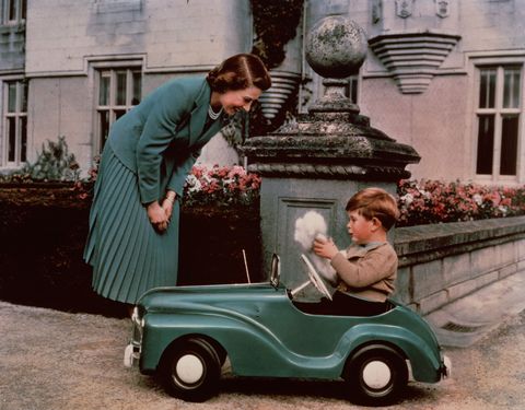 princess elizabeth stands and watches her 3 year old son prince charles who is sitting in a toy car that is green and holding a white ball of fluff