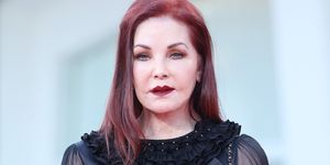 priscilla presley looks at the camera standing, she wears a black blouse with a ruffled collar and sheer long sleeves