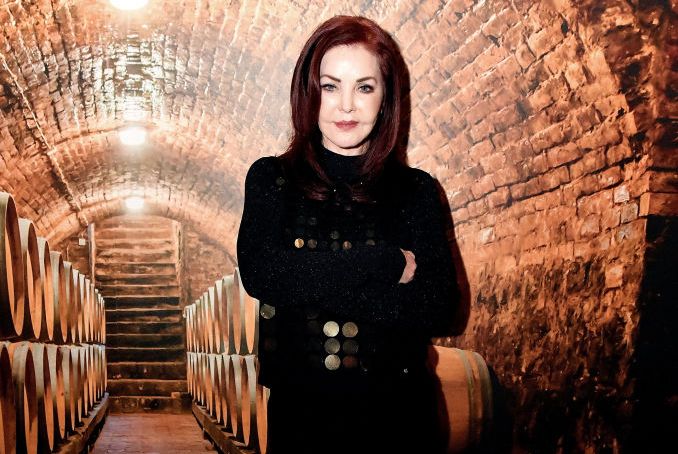 priscilla presley stands in front of several wine barrels in a stone arched cellar, she looks at a camera with a slight smile, her arms are crossed and she is wearing a black outfit