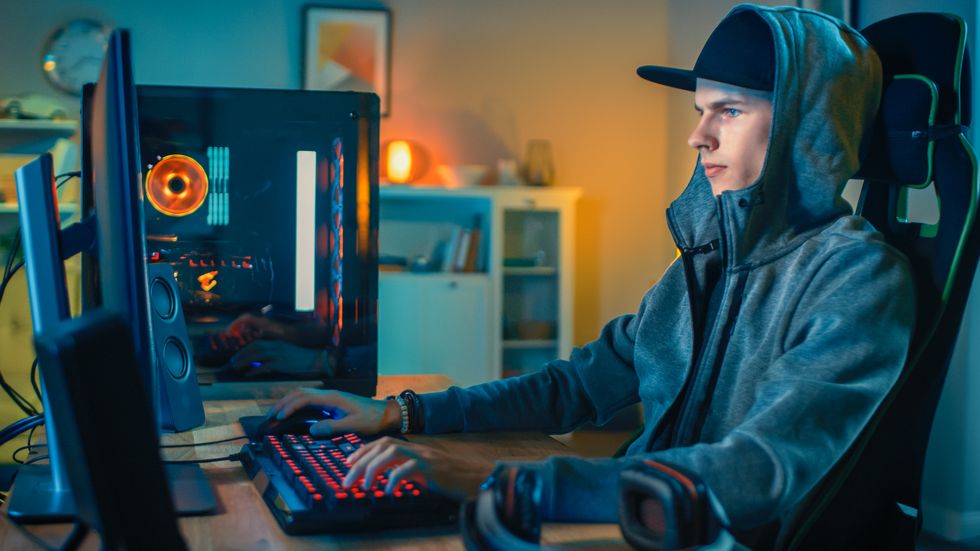 professional gamer or streamer playing first person shooter online video game on his cool personal computer young man is wearing a cap and hood room and pc have colorful neon led lights cozy evening at home