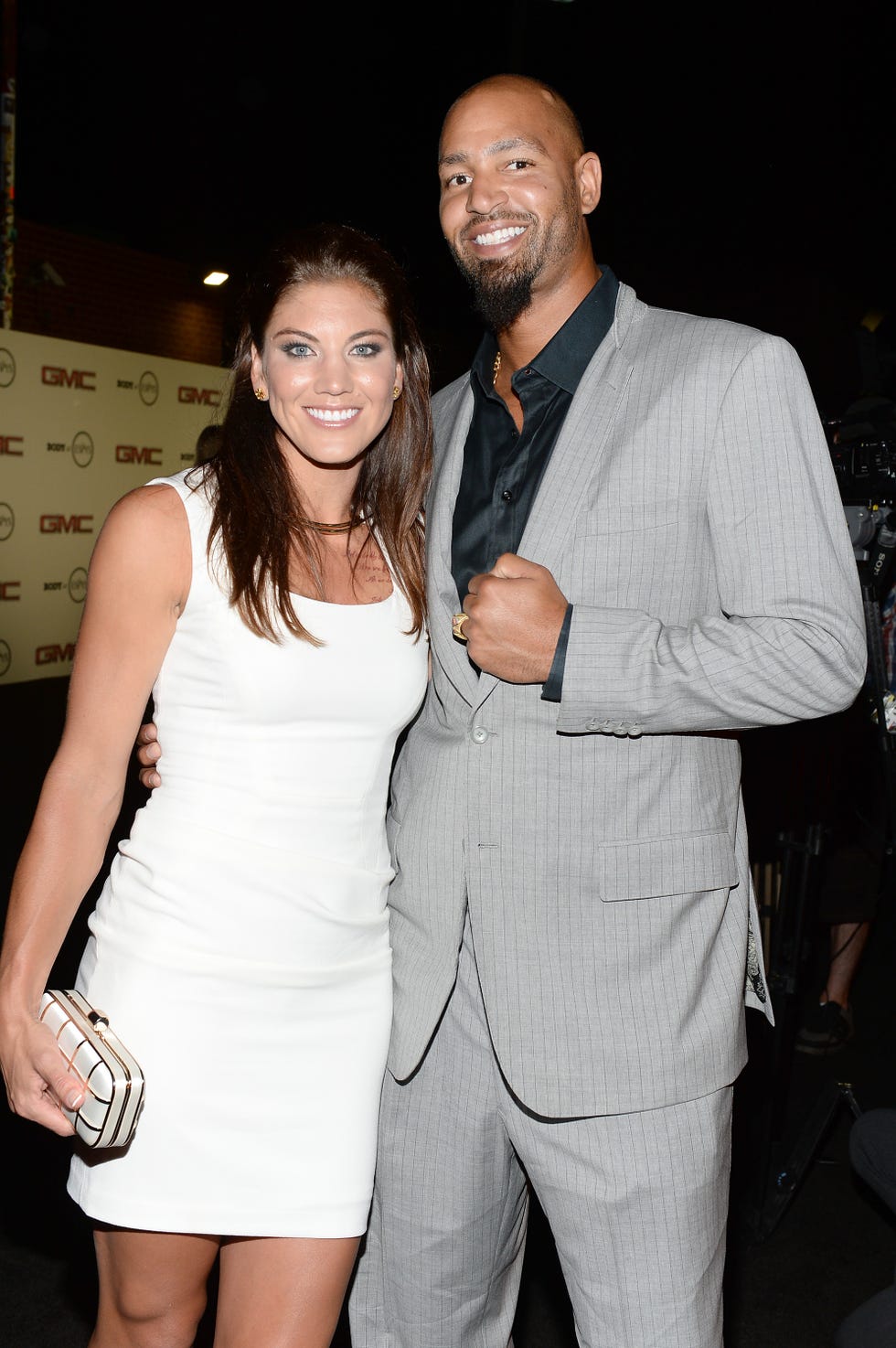 hope solo and jerramy stevens stand together and smile for a photo, she wears a white sleeveless dress, he wears a gray pinstriped suit jacket over a black collared shirt