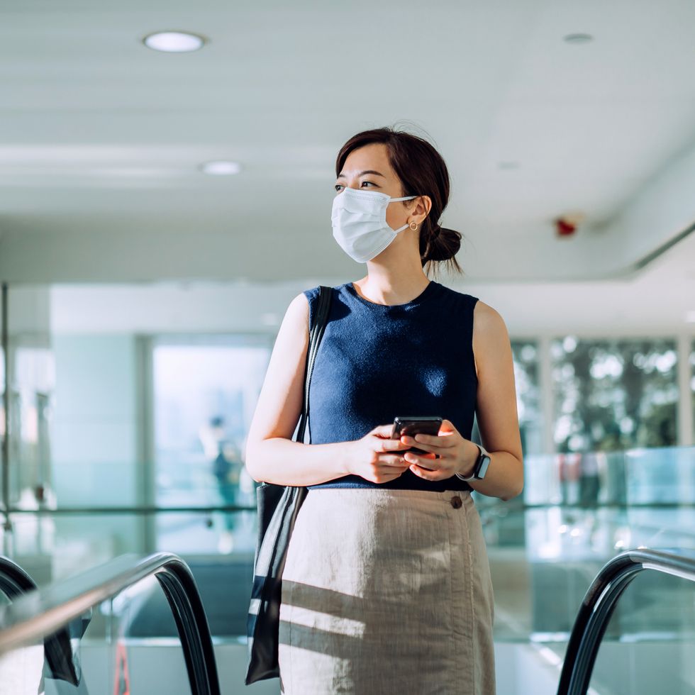 professional young asian businesswoman with protective face mask commuting to work in the morning, holding smartphone on hand while riding on the escalator in an office building