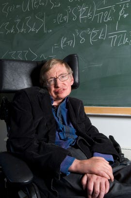 stephen hawking smiles at the camera while sitting in his wheelchair in front of a green chalkboard with written equations, he wears a dark suit jacket and blue collared shirt with white pinstripes