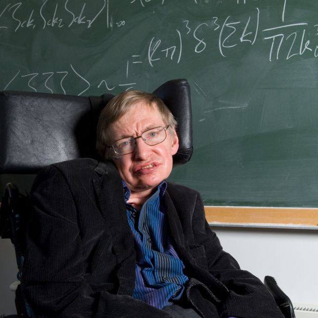 stephen hawking smiles at the camera while sitting in his wheelchair in front of a green chalkboard with written equations, he wears a dark suit jacket and blue collared shirt with white pinstripes