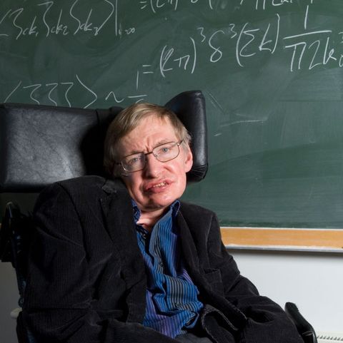 stephen hawking smiles at the camera while sitting in his wheelchair in front of a green chalkboard with written equations, he wears a dark suit jacket and blue collared shirt with white pinstripes