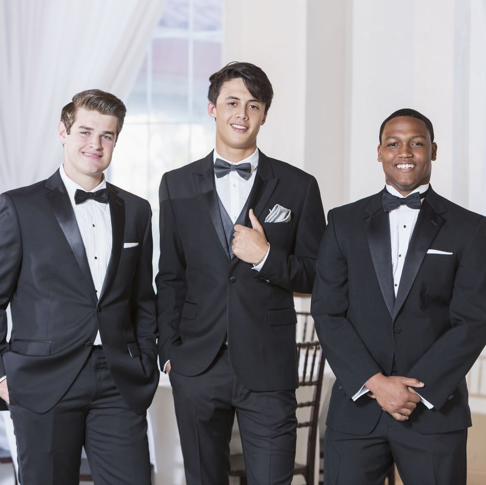 a group of three multi ethnic young men wearing tuxedos, standing in a row, smiling at the camera they are at a party, prom, a wedding or some other special formal event