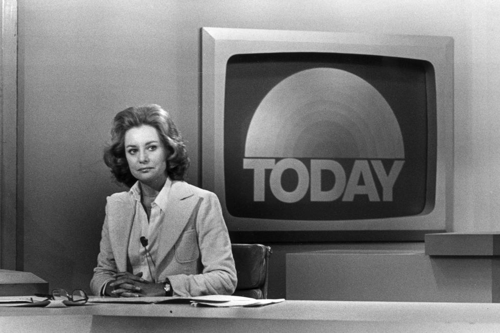 barbara walters sits at a news desk with her hands clasped on top of papers, she is sitting in front of a framed monitor that shows the today show logo