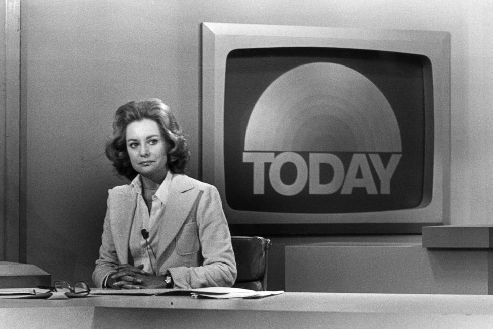 barbara walters sits at a news desk with her hands clasped on top of papers, she is sitting in front of a framed monitor that shows the today show logo