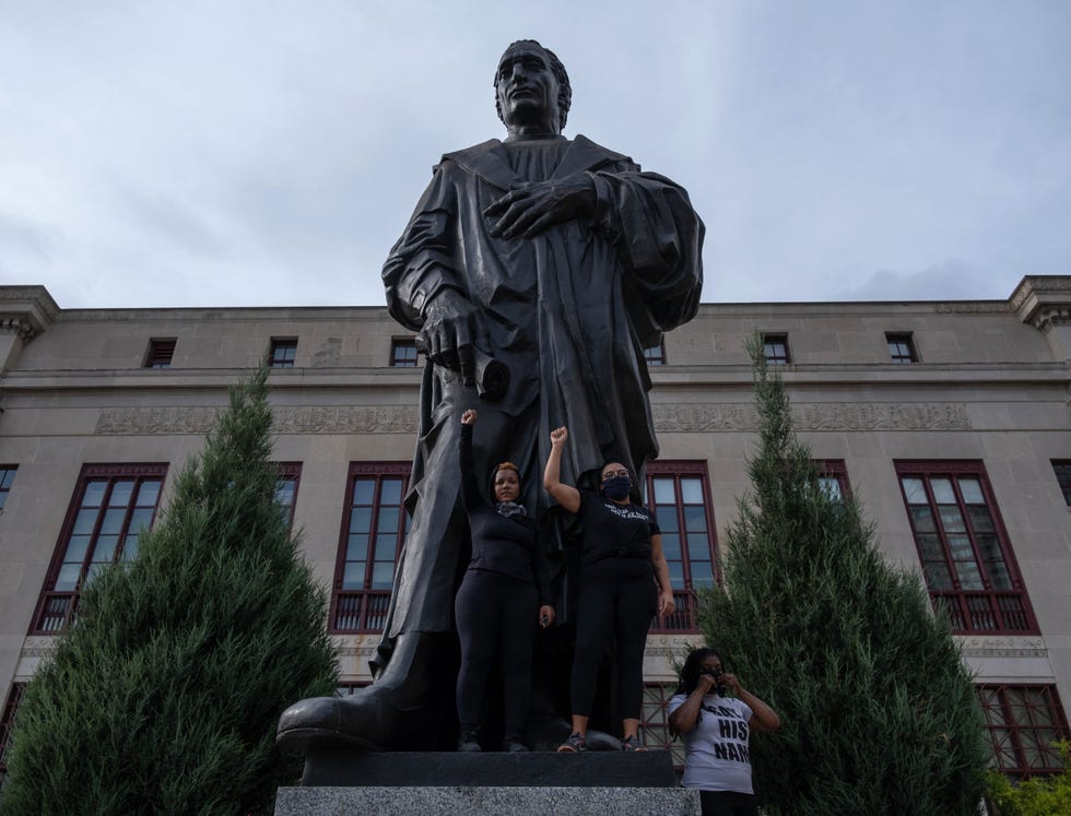 Columbus, Ohio has moved away from celebrating namesake Christopher Columbus two protestors holding their arm in the air in front of a metal statue of christopher columbus